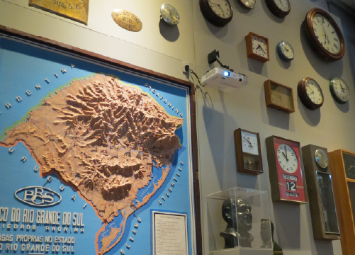 Foto de parede com diversos rel�gios e o mapa do relevo do Estado do Rio Grande do Sul em relevo.